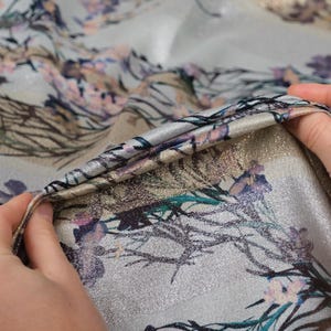 May include: Close-up of a silver fabric with a floral print. The fabric has a subtle shimmer and a soft, flowing texture.