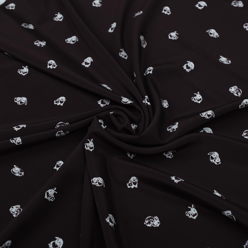 May include: Black fabric with a repeating pattern of white bunny illustrations. The fabric is draped, showing folds and shadows. The bunnies are depicted in a simple, cartoonish style, scattered across the surface.