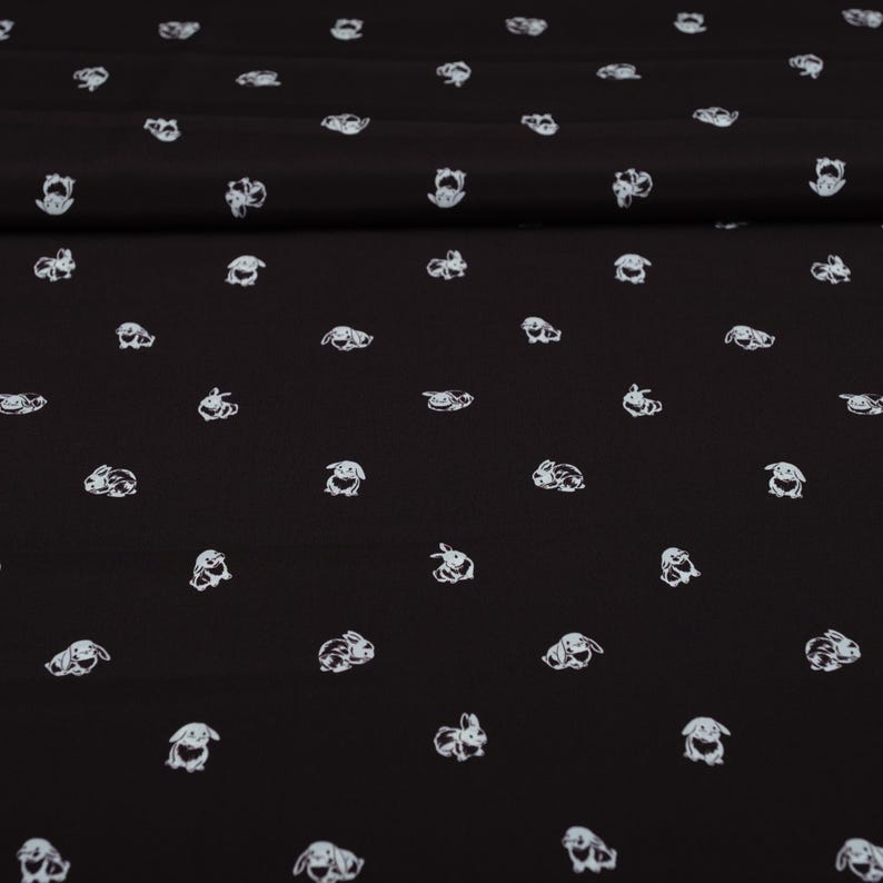 May include: Black fabric with a repeating pattern of white bunny illustrations. The bunnies are depicted in various poses, creating a playful and whimsical design. The fabric appears to be soft and suitable for various sewing projects.