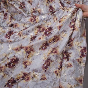 May include: A white fabric with a floral pattern in shades of gold and burgundy. The fabric has a subtle sheen and a soft, flowing texture.