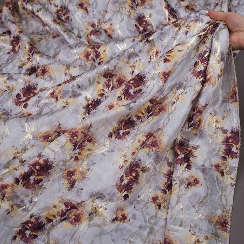 May include: A white fabric with a floral pattern in shades of gold and burgundy. The fabric has a subtle sheen and a soft, flowing texture.