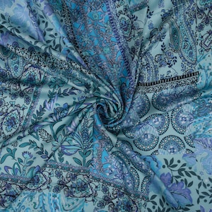 Op de afbeelding: Een close-up van een zijdeachtige, bedrukte stof in tinten blauw en teal. De stof heeft ingewikkelde bloemen- en paisley-ontwerpen, met een wervelende, getextureerde uitstraling. De algehele esthetiek doet denken aan een sjaal of omslagdoek.