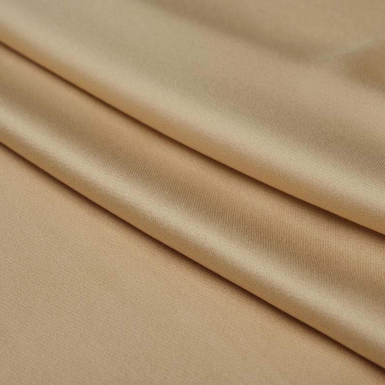 May include: Close-up of a folded fabric in a light tan color. The fabric has a smooth, slightly shiny texture, with visible folds and shadows that add depth. The overall appearance is soft and flowing.