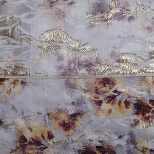 May include: A close-up of a light gray fabric with a floral pattern. The fabric has a subtle shimmer and a textured surface. The flowers are a mix of purple and yellow.