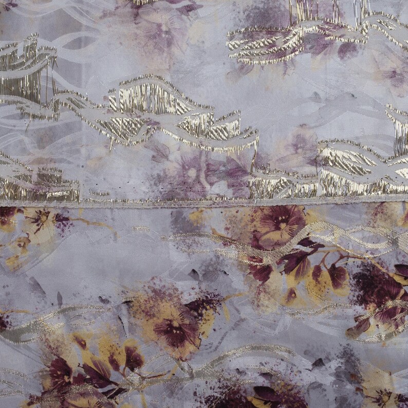 May include: A close-up of a light gray fabric with a floral pattern. The fabric has a subtle shimmer and a textured surface. The flowers are a mix of purple and yellow.