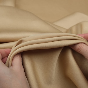 May include: Close-up of a beige fabric with soft folds. The fabric is held in the hands, showcasing its texture and drape. The color is a warm, neutral tone, suitable for various textile projects.