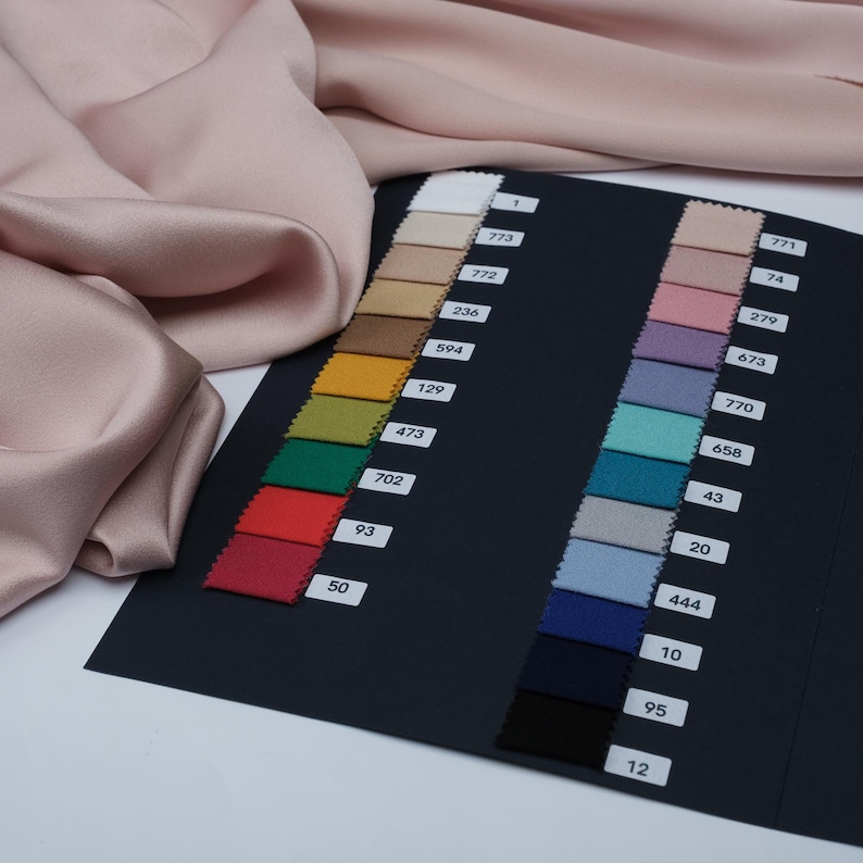 May include: A swatch card with fabric samples in various colors, including pink, beige, and blue, is displayed next to a piece of draped, silky fabric. Each swatch is labeled with a number. The fabric appears to be satin or a similar material.