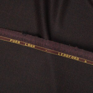 May include: Close-up of a dark brown fabric with a subtle textured pattern. The fabric has a burgundy colored border with the text "PURA LANA - LEADFORD &" in gold lettering.