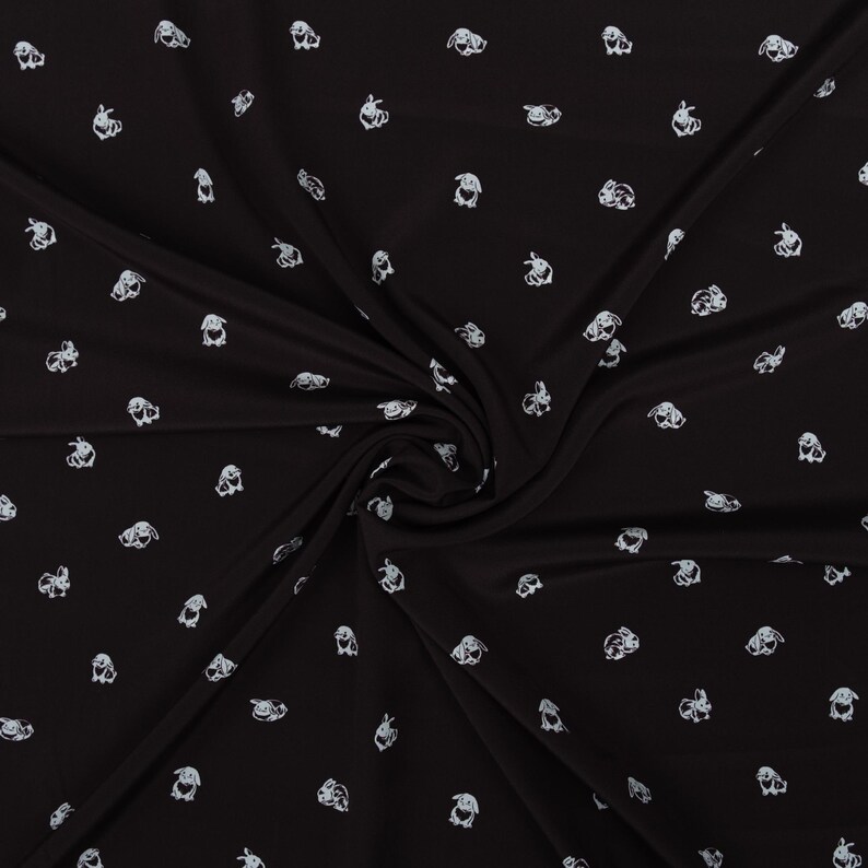 May include: Black fabric with a repeating pattern of white bunny illustrations. The bunnies are depicted in various poses and orientations, creating a playful and whimsical design. The fabric is draped, showing folds and shadows.