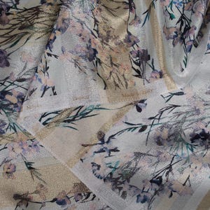 May include: A close-up of a white fabric with a gold and silver metallic sheen. The fabric has a floral print with pink, blue, and green tones. The fabric has a label that reads "Made in Italy".