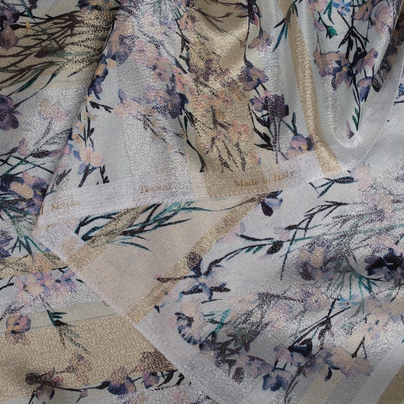 May include: A close-up of a white fabric with a gold and silver metallic sheen. The fabric has a floral print with pink, blue, and green tones. The fabric has a label that reads "Made in Italy".