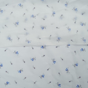 May include: White fabric with a delicate pattern of small blue flowers and tiny dark green accents. The floral design is evenly distributed across the fabric, creating a soft, airy aesthetic. The fabric appears to be lightweight and suitable for various sewing projects.