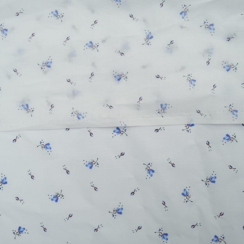 May include: White fabric with a delicate pattern of small blue flowers and tiny dark green accents. The floral design is evenly distributed across the fabric, creating a soft, airy aesthetic. The fabric appears to be lightweight and suitable for various sewing projects.