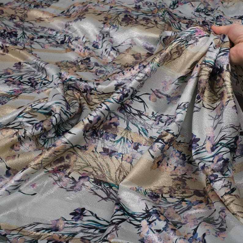 May include: Silver fabric with a floral print in shades of pink, blue, and gold. The fabric has a subtle shimmer and a soft, flowing texture.