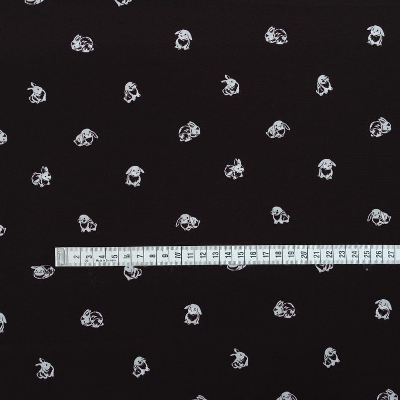 May include: Black fabric with a repeating pattern of white bunny illustrations. A ruler is placed across the bottom of the fabric, showing measurements in inches. The bunnies are in various poses.