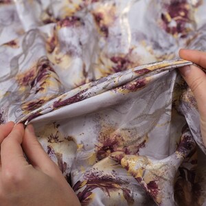 May include: Close-up of a soft, white fabric with a floral print in shades of purple, gold, and brown. The fabric is textured and has a subtle sheen.