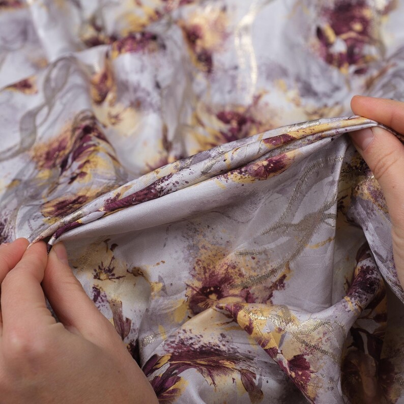May include: Close-up of a soft, white fabric with a floral print in shades of purple, gold, and brown. The fabric is textured and has a subtle sheen.
