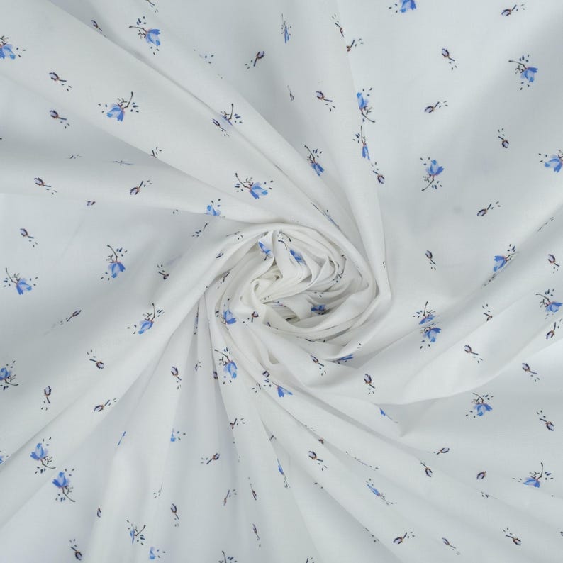May include: White fabric with a repeating pattern of small blue floral sprigs. The fabric is gathered in a swirl, showing the texture and the delicate design. The flowers have blue petals and dark stems.