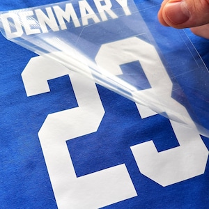 May include: A bright blue fabric surface featuring a white heat transfer vinyl design of the number 23 and the word DENMARK. A clear plastic carrier sheet is being peeled away from the white vinyl lettering and numbering on the blue textile.