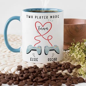May include: White coffee mug with a blue handle and rim, featuring the text "TWO PLAYER MODE Forever" and two game controllers. The mug is surrounded by coffee beans and a wooden bowl.