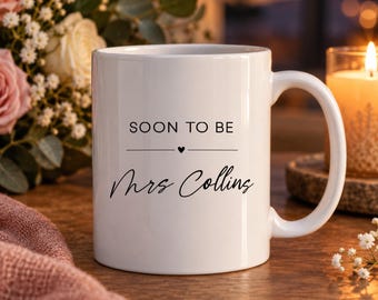 May include: A white ceramic mug featuring the black printed text SOON TO BE followed by a small heart icon and the name Mrs Collins in a cursive script. The mug has a smooth finish and a curved handle, presented as a functional drinkware item.