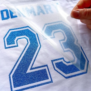 May include: A white t-shirt featuring a blue glitter heat transfer vinyl design with the number 23 and the partial text DENMARK. A clear plastic carrier sheet is being peeled away from the blue glitter number, revealing the textured finish.