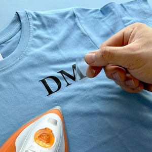 May include: A light blue t-shirt featuring the black letters DM being applied with a heat transfer process. An orange and white iron is positioned at the bottom, while a clear plastic film is being peeled away from the black vinyl lettering.
