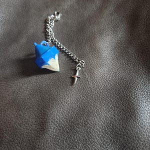 May include: A geometric pendant with a blue and white design, suspended from a silver chain. A small silver cross charm dangles below. The pendant is a unique accessory.