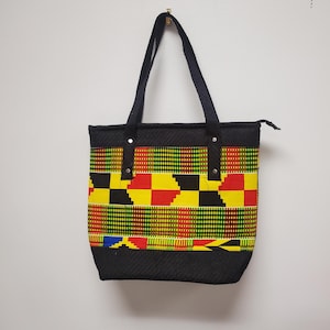 Könnte beinhalten: Schwarze Tragetasche mit Reißverschluss und schwarzen Henkeln. Die Tasche ist aus einem gewebten Stoff mit einem bunten geometrischen Muster in den Farben Gelb, Rot, Grün und Schwarz.