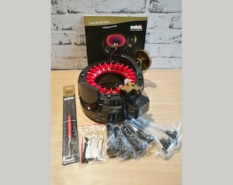 May include: A black and red circular knitting machine with a gold knob and a clear plastic box containing knitting needles. The machine is labeled "addi" and the box is labeled "addi Premium".