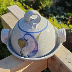 May include: A ceramic butter dish with a lid and handles. The dish is off-white with blue and purple floral designs. The lid has a loop handle. The dish sits on a wooden surface.