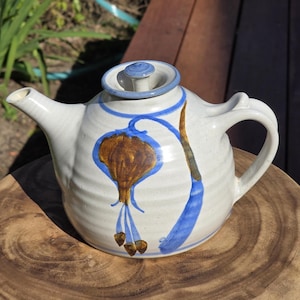 May include: Cream-coloured ceramic teapot with a blue and brown floral pattern. The teapot features a curved spout, a handle, and a lid with a blue knob. The floral design includes a brown bulb shape with blue accents.