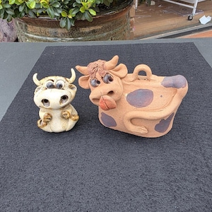 May include: Two ceramic cow figurines. One is off-white with googly eyes and gold accents. The other is brown with purple spots and a tongue sticking out. Both are whimsical and decorative.