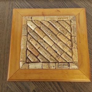 May include: Square wooden coaster with a frame, featuring a mosaic of wine corks arranged diagonally. The corks, with printed text, are set within a light brown wooden frame, creating a rustic, decorative item for drinks.