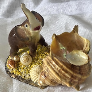 May include: A brown ceramic elephant figurine with a raised trunk standing on a base covered in yellow gravel, small white and brown seashells, and a large, spiraled, tan-coloured conch shell positioned next to the elephant on the base.