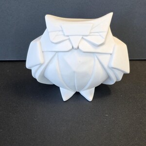 May include: A white origami owl figurine with a geometric design. The owl has a bow tie and a folded body, with pointed feet. The figurine is set against a dark background.