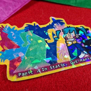 May include: A holographic sticker with a cartoon character design. The sticker has a yellow border and text that reads "Panic In Static. Out-Manic." The background is red with colored pencils and paper.