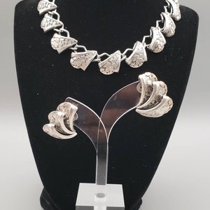 May include: A silver necklace and earrings set with a geometric design. The necklace is made of linked, textured, fan-shaped pieces. The earrings are also fan-shaped and textured.