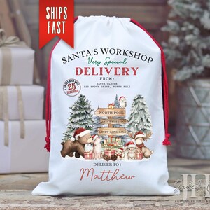 May include: White Christmas gift bag with red drawstring and trim. The bag features a festive illustration of Santa's Workshop with woodland creatures and a North Pole sign. Text includes "Santa's Workshop" and "Delivery to Matthew."