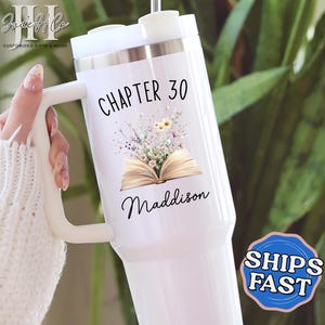 May include: White insulated tumbler with a handle, featuring the text "CHAPTER 30" above an illustration of an open book and flowers. The name "Maddison" is written below the illustration. The tumbler has a silver rim and a white lid.
