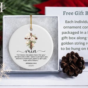 May include: White ceramic Christmas ornament with a floral cross design and the name "mia". The ornament includes a gold string for hanging and is packaged in a gift box. The text "For I know the plans I have for you" is also included.