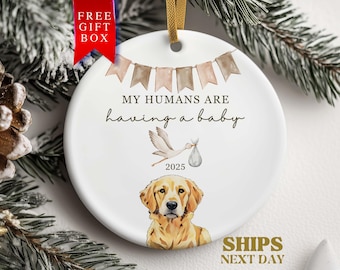 May include: White ceramic Christmas ornament with a gold ribbon. The ornament features the text "MY HUMANS ARE having a baby" with a stork carrying a baby and a golden retriever. The year 2025 is also displayed.