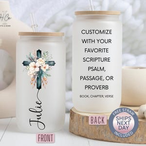 May include: Two frosted glass tumblers with a wooden lid and a straw. The tumbler on the left has a watercolor floral cross design and the name "Julie" written below. The tumbler on the right has the text "Customize with your favorite scripture psalm, passage, or proverb. Book, chapter, verse."