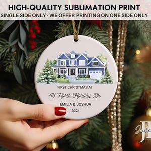 May include: A white ceramic Christmas ornament with a watercolor illustration of a blue house with white trim. The text "First Christmas at 48 North Holiday Dr Emilia & Joshua 2024" is printed on the ornament.