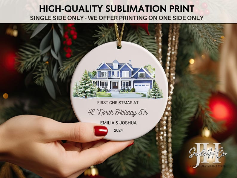 May include: A white ceramic Christmas ornament with a watercolor illustration of a blue house with white trim. The text "First Christmas at 48 North Holiday Dr Emilia & Joshua 2024" is printed on the ornament.