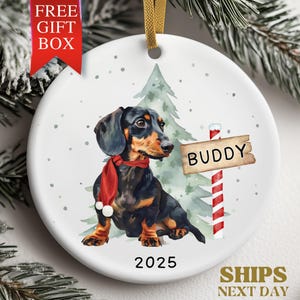May include: A round Christmas ornament featuring a dachshund wearing a red scarf, with a sign that says "Buddy". The ornament has a gold ribbon for hanging and the year 2025 is printed on the bottom. The words "Free Gift Box" are printed on a red banner.