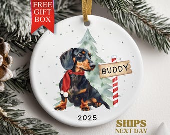 May include: A round Christmas ornament featuring a dachshund wearing a red scarf, with a sign that says "Buddy". The ornament has a gold ribbon for hanging and the year 2025 is printed on the bottom. The words "Free Gift Box" are printed on a red banner.