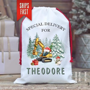 May include: White drawstring Christmas gift bag with red trim and drawstrings. The bag features a festive design with a yellow excavator wearing a Santa hat, a snowman, and the text "SPECIAL DELIVERY FOR THEODORE".