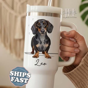 May include: White insulated tumbler featuring a black, brown, and tan dachshund illustration. The tumbler has a handle and a straw. The name "Leo" is written below the dog. The tumbler has a white lid and a silver band.