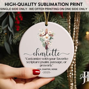 May include: A round, white ceramic ornament with a floral cross design and the name "Charlotte" in script. The ornament includes the text "Customize with your favorite scripture psalm, passage, or proverb."